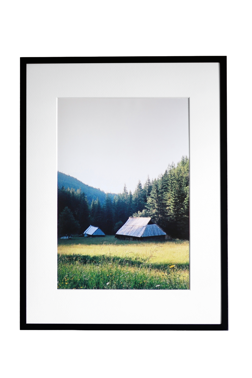 A framed landscape photograph print featuring a mountain, pine forest, and two small tents in a grassy meadow, presented in a black frame with a white mat against a white background.