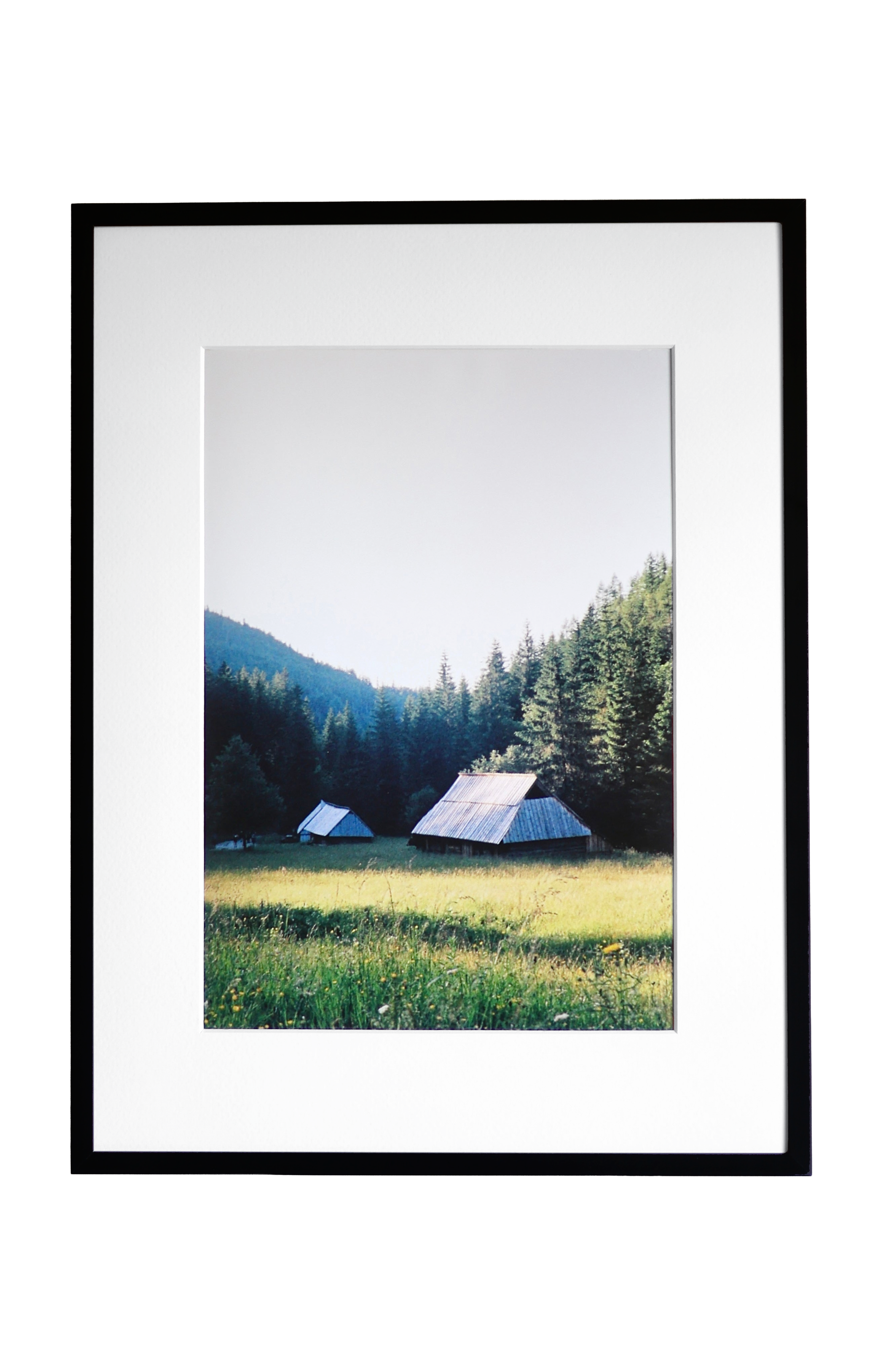 A framed landscape photograph print featuring a mountain, pine forest, and two small tents in a grassy meadow, presented in a black frame with a white mat against a white background.