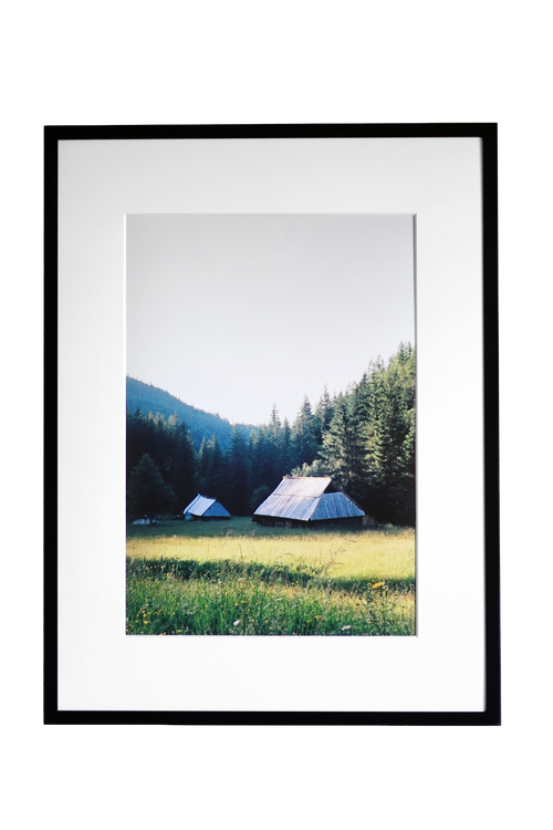 A framed landscape photograph print featuring a mountain, pine forest, and two small tents in a grassy meadow, presented in a black frame with a white mat against a white background.
